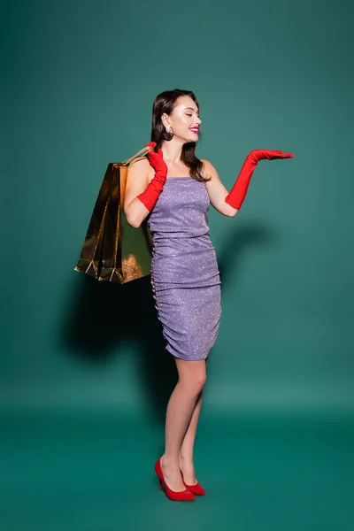 Pleine longueur de femme heureuse en gants rouges et robe violette tenant des sacs à provisions tout en pointant avec la main sur le vert — Photo de stock