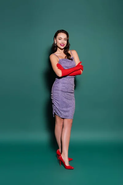 Full length of brunette young woman in red gloves and purple dress smiling while posing with crossed arms on green — Stock Photo