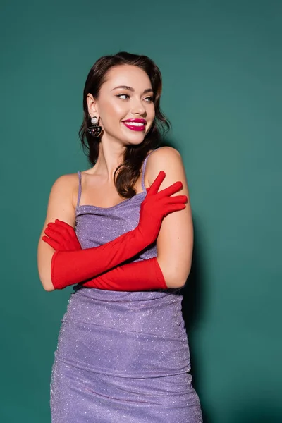 Young woman in red gloves and purple dress smiling while posing with crossed arms on green — Stock Photo