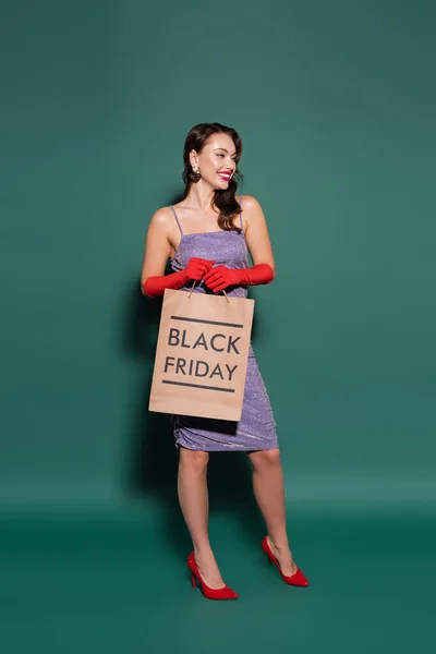 Happy young woman in red gloves and purple dress holding shopping bag with black friday lettering on green — Stock Photo