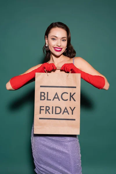 Jeune femme souriante en gants rouges et robe violette tenant sac à provisions avec lettrage noir vendredi sur vert — Photo de stock