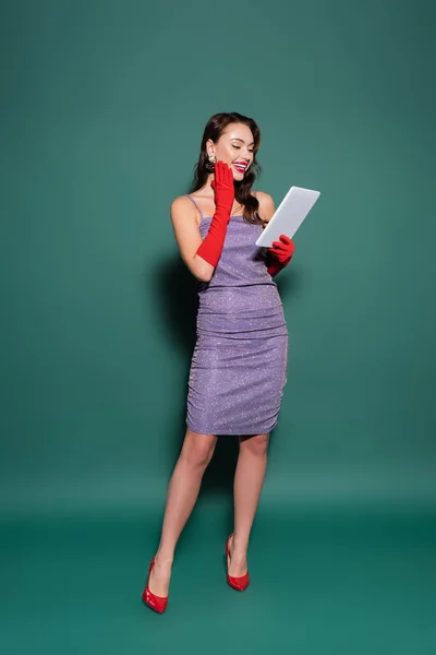 Longitud completa de la joven feliz en vestido púrpura y guantes rojos usando tableta digital en verde - foto de stock