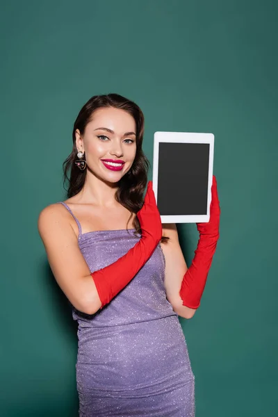 Mujer joven feliz en vestido púrpura y guantes que sostienen la tableta digital con pantalla en blanco en verde - foto de stock