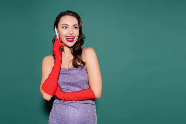 Joyeuse jeune femme en robe violette et gants parlant sur smartphone sur vert — Photo de stock