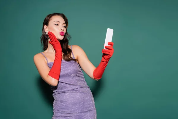 Young woman in purple dress and gloves pouting lips while taking selfie on smartphone on green — Stock Photo