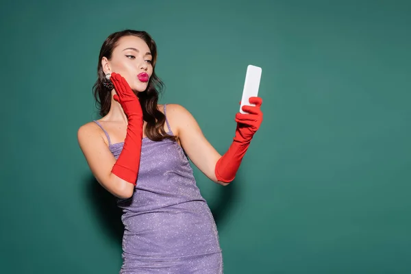 Jeune femme en robe violette et gants boudant les lèvres tout en prenant selfie sur smartphone sur vert — Photo de stock