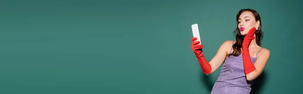 Mujer joven en vestido púrpura y guantes haciendo pucheros labios mientras toma selfie en el teléfono inteligente en verde, bandera - foto de stock