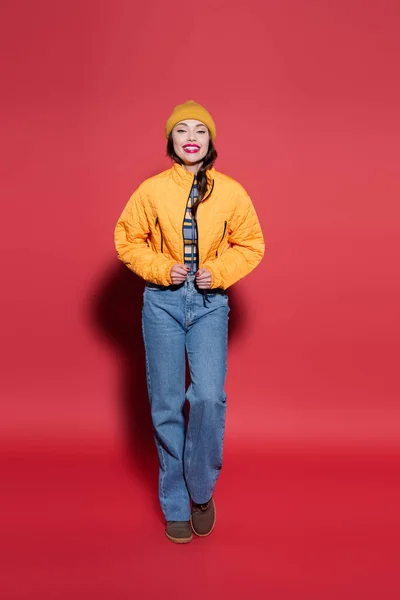 Longitud completa de la joven feliz en gorro sombrero ajustando chaqueta hinchable naranja y sonriendo en rojo - foto de stock