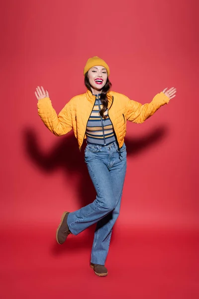 Comprimento total de feliz jovem mulher em chapéu gorro e jaqueta de puffer laranja se divertindo no vermelho — Fotografia de Stock