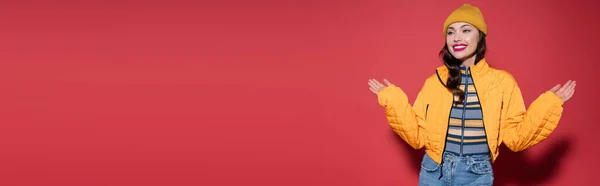 Feliz joven mujer en gorro sombrero y chaqueta hinchable naranja y sonriendo mientras gesticulaba en rojo, bandera - foto de stock