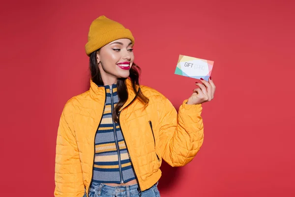 Happy young woman in beanie hat and orange puffer jacket holding gift card on red — Stock Photo