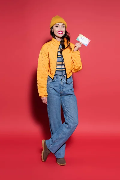 Pleine longueur de jeune femme heureuse en bonnet chapeau et veste gonflante orange tenant carte cadeau sur rouge — Photo de stock