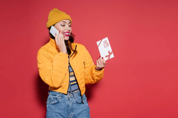 Femme heureuse en bonnet chapeau et veste gonflante orange tenant carte cadeau tout en parlant sur smartphone sur rouge — Photo de stock