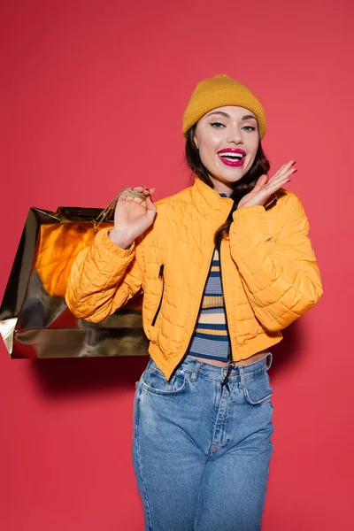 Excited young woman in orange beanie hat and puffer jacket holding shopping bag on red — Stock Photo