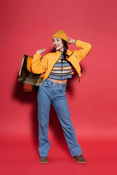 Piena lunghezza di giovane donna positiva in cappello di berretto arancione e giacca di puffer che tiene la shopping bag sul rosso — Foto stock