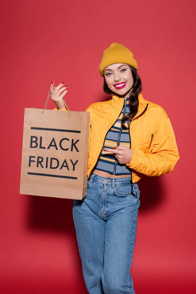 Heureuse jeune femme en bonnet orange chapeau et veste soufflante pointant vers le sac à provisions avec lettrage noir vendredi sur rouge — Photo de stock