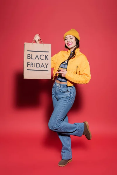 In voller Länge glückliche junge Frau mit Mütze und Pufferjacke zeigt auf Einkaufstasche mit schwarzem Freitag-Schriftzug auf rot — Stockfoto