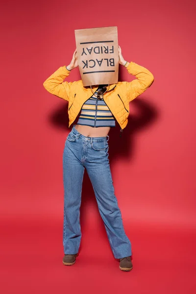 Full length of woman in orange puffer jacket with black friday paper bag on head standing on red — Stock Photo