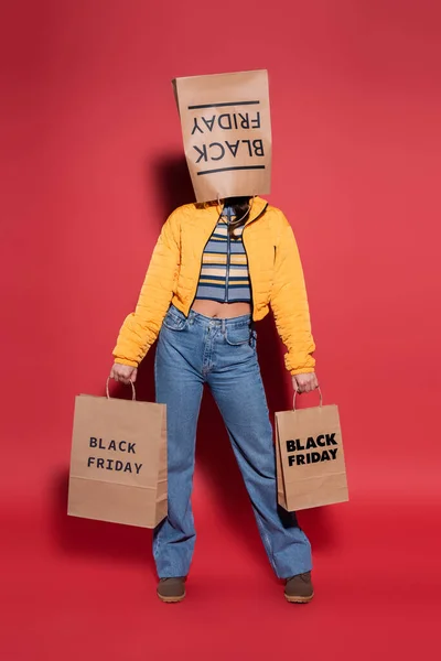 Volle Länge der Frau in orangefarbener Pufferjacke mit schwarzer Freitagspapiertasche auf dem Kopf stehend mit Einkaufstaschen auf rotem Grund — Stockfoto
