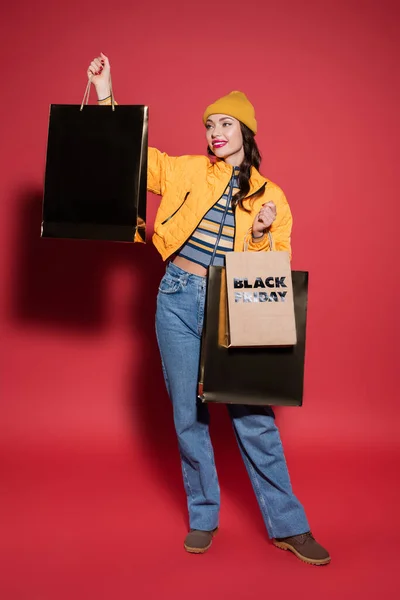 Full length of cheerful woman in beanie hat and puffer jacket holding shopping bags with black friday lettering on red — Stock Photo
