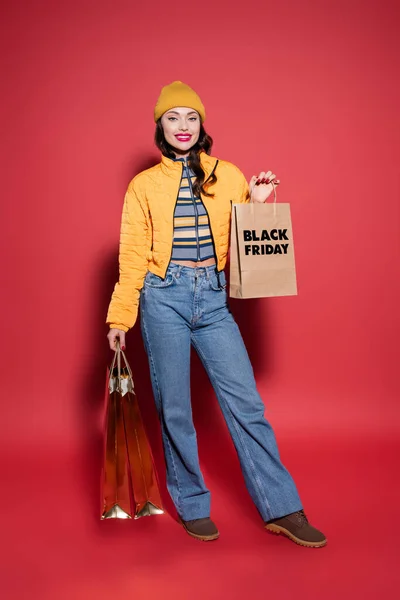 Full length of smiling woman in beanie hat and puffer jacket holding shopping bags with black friday lettering on red — Stock Photo