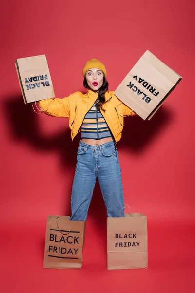 Femme choquée en bonnet chapeau et veste gonflable avec sacs à provisions vendredi noir sur les mains debout sur rouge — Photo de stock