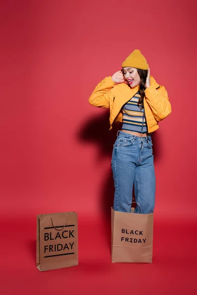 In voller Länge staunende Frau mit Mütze, die in einer Einkaufstasche mit schwarzem Freitag-Schriftzug auf Rot steht — Stockfoto