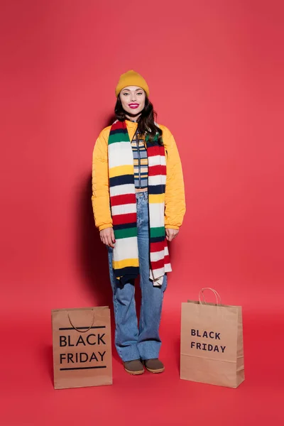 In voller Länge lächelnde Frau mit Mütze und Schal, die neben Einkaufstüten mit schwarzem Freitag-Schriftzug auf Rot steht — Stockfoto