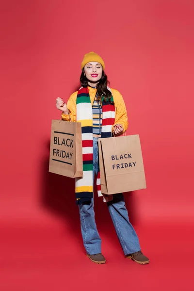 In voller Länge glückliche Frau mit Mütze und Schal, die Einkaufstaschen mit schwarzem Freitag-Schriftzug auf Rot hält — Stockfoto