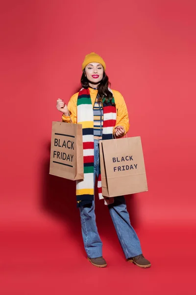 Pleine longueur de femme heureuse en bonnet chapeau et écharpe tenant des sacs à provisions avec lettrage noir vendredi sur rouge — Photo de stock