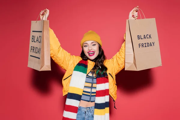 Amazed woman in beanie hat and scarf holding shopping bags with black friday lettering on red — Stock Photo