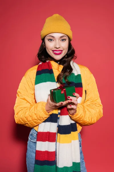 Happy woman in beanie hat and scarf holding wrapped gift box on red — Stock Photo