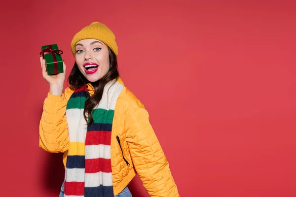 Excited woman in beanie hat and scarf holding wrapped gift box on red — Stock Photo