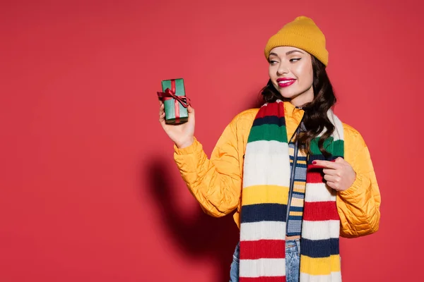 Felice donna in cappello beanie e sciarpa che punta con il dito a scatola regalo avvolto su rosso — Foto stock