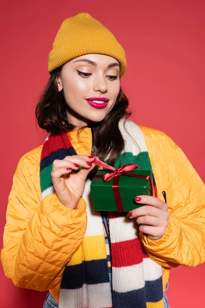 Улыбающаяся молодая женщина в шапочке и шарфе, смотрящая на завернутую подарочную коробку на красном — стоковое фото