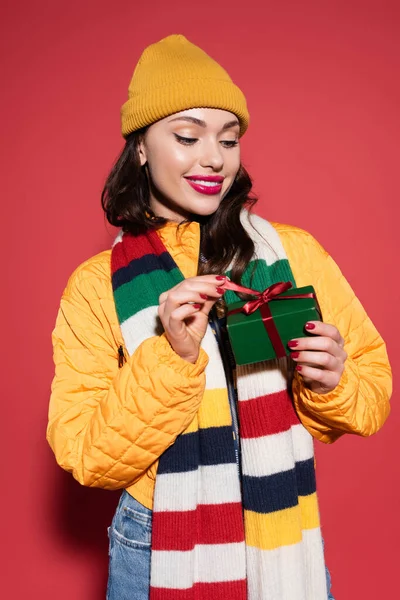 Femme positive en bonnet chapeau et écharpe tenant boîte cadeau enveloppée sur rouge — Photo de stock