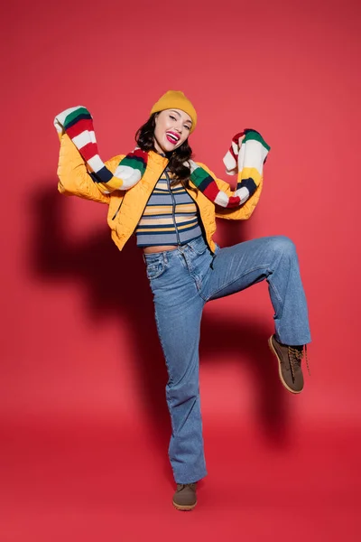 Pleine longueur de femme positive en bonnet chapeau et veste de bouffée orange tenant foulard dépouillé sur rouge — Photo de stock