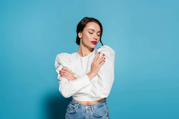 Encantadora joven en blusa blanca posando con los brazos cruzados y los ojos cerrados aislados en azul - foto de stock