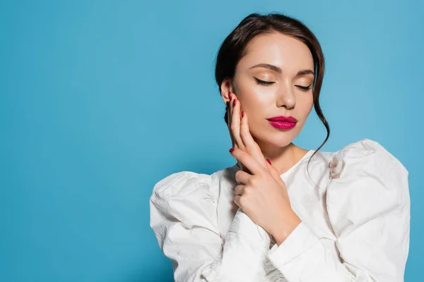 Portrait of sensual young woman in white blouse posing with closed eyes and touching face isolated on blue — Stock Photo