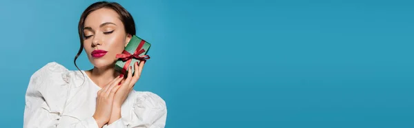 Brunette woman in white blouse holding wrapped present and posing with closed eyes isolated on blue, banner — Stock Photo