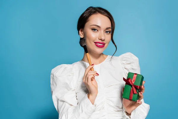 Happy young woman in white blouse holding wrapped present and cosmetic brush isolated on blue — Stock Photo