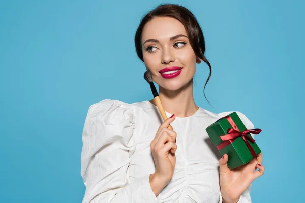 Fröhliche junge Frau in weißer Bluse mit eingewickeltem Geschenk und Kosmetikpinsel isoliert auf blauem Grund — Stockfoto