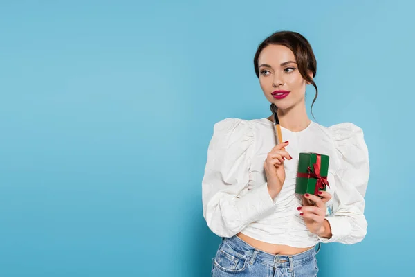 Verträumte junge Frau in weißer Bluse mit eingewickeltem Geschenk und Kosmetikpinsel auf blauem Grund — Stockfoto