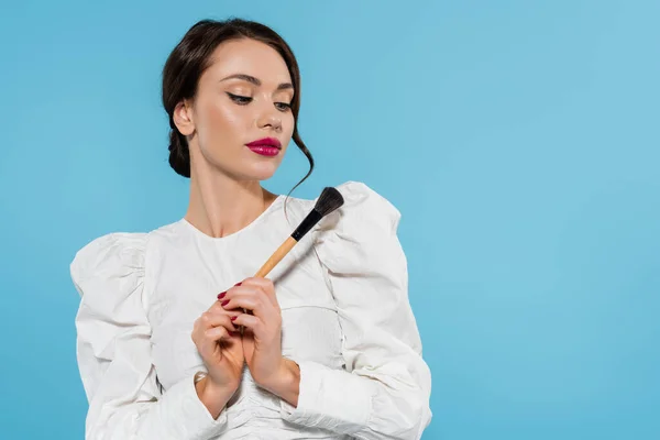 Jovem morena em blusa branca segurando escova cosmética isolada em azul — Fotografia de Stock