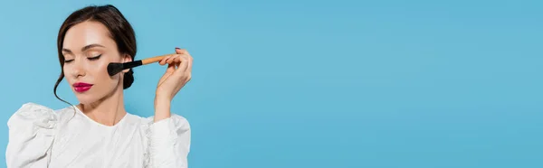 Brunette young woman in white blouse holding cosmetic brush near cheek isolated on blue, banner — Stock Photo