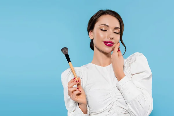 Alegre joven en blusa blanca sosteniendo cepillo cosmético y tocando mejilla aislada en azul — Stock Photo