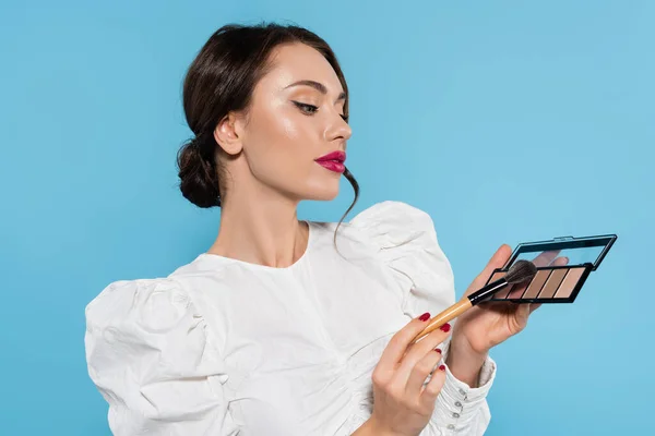 Brunette young woman in white blouse holding cosmetic brush and eye shadow palette isolated on blue — Stock Photo