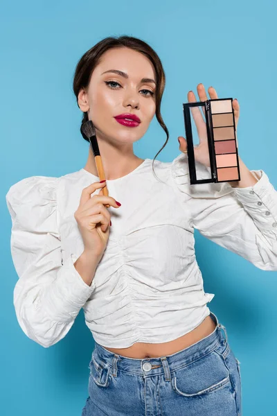 Young woman in white blouse holding cosmetic brush and eye shadow palette isolated on blue — Stock Photo