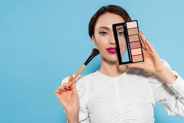 Morena mujer joven en blusa blanca con cepillo cosmético y sombra de ojos paleta cerca de la cara aislada en azul - foto de stock