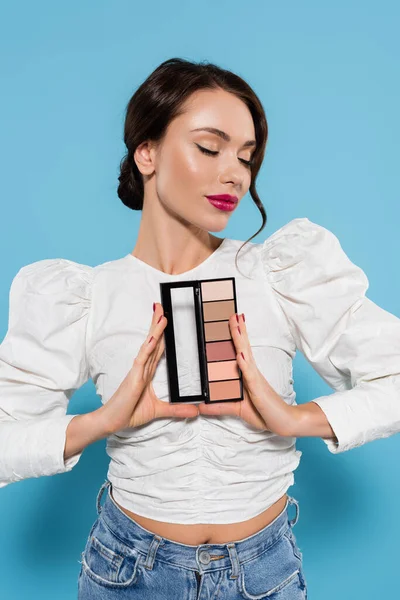 Morena joven en blusa blanca sosteniendo la paleta de sombras de los ojos y de pie con los ojos cerrados aislados en azul - foto de stock